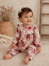 Smiling toddler in floral pink romper, sitting on a textured rug with a natural background.