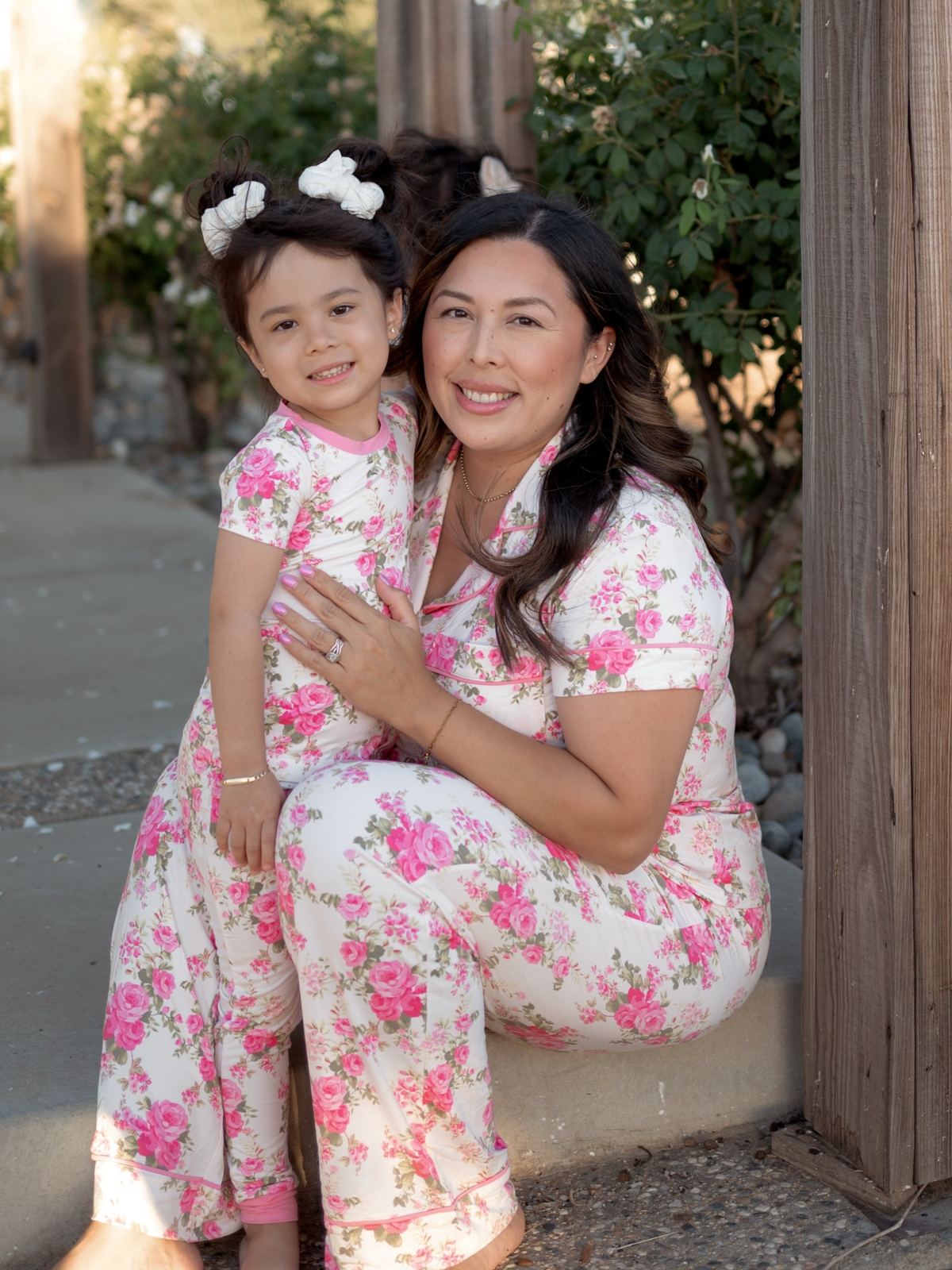 Mother and daughter in matching floral pajamas, smiling together outdoors near wooden posts and greenery.