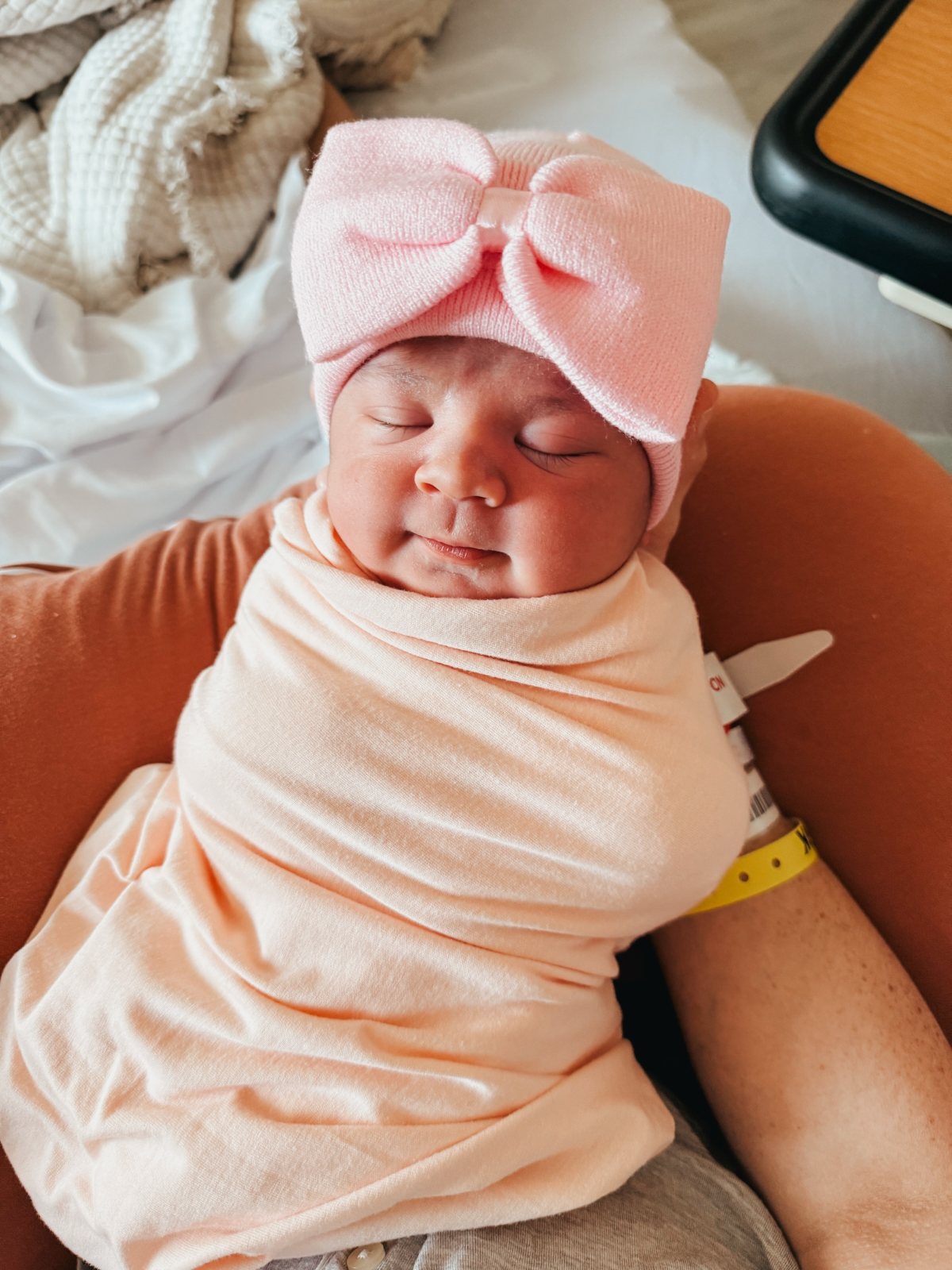 Newborn baby with a pink bow headband, peacefully swaddled, resting in a cozy setting.