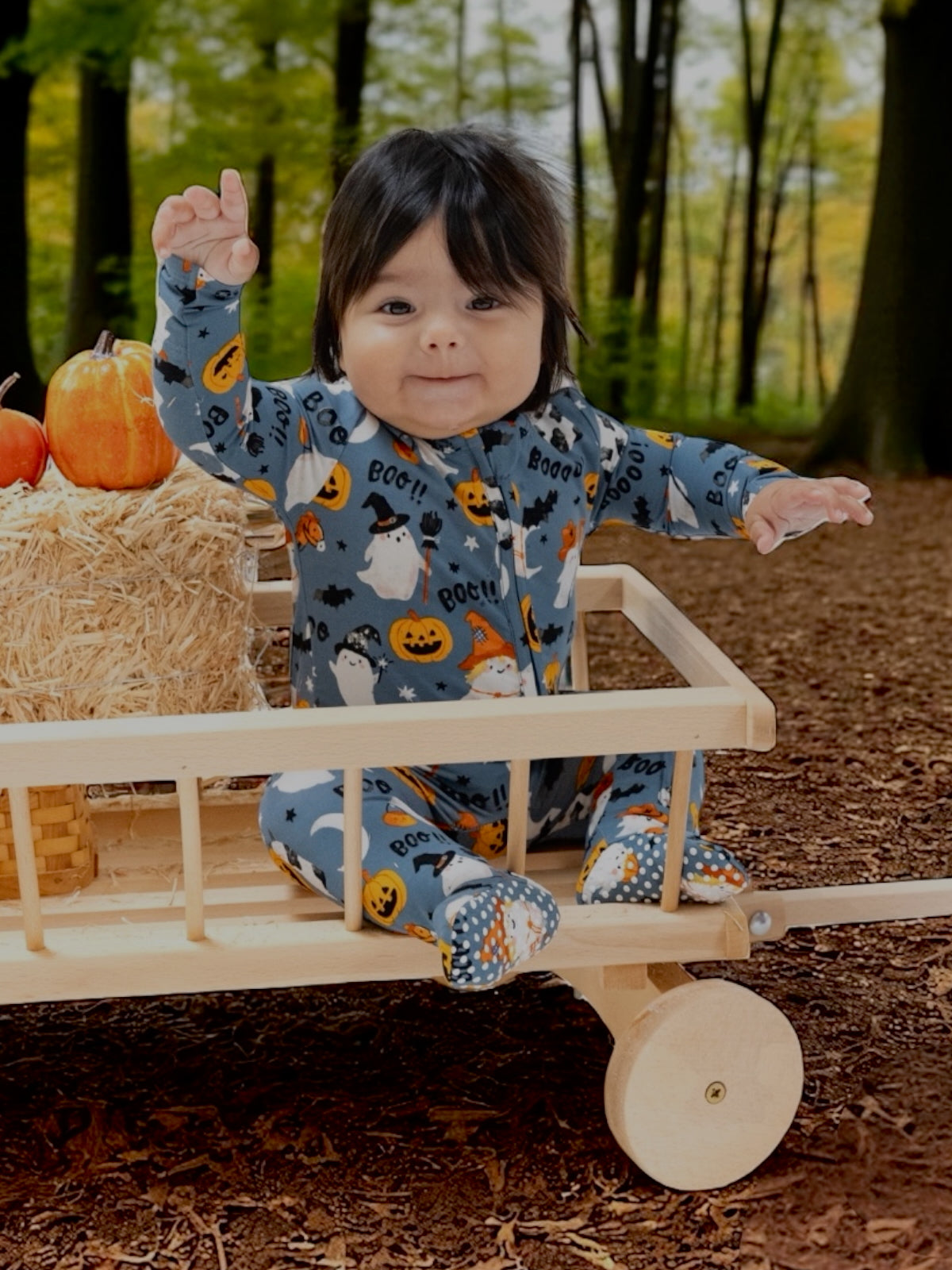 Infant in Halloween-themed pajamas sits in a wooden wagon with pumpkins and hay in a wooded area.
