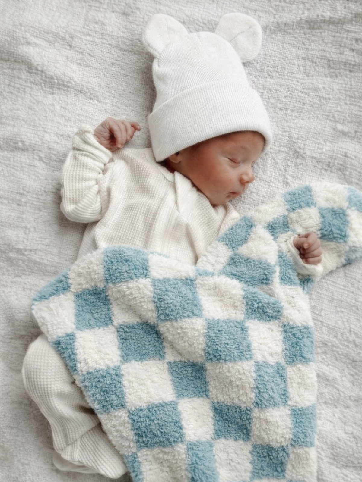 Newborn baby wearing a white bear hat, peacefully sleeping under a blue and white checkered blanket on a textured surface.