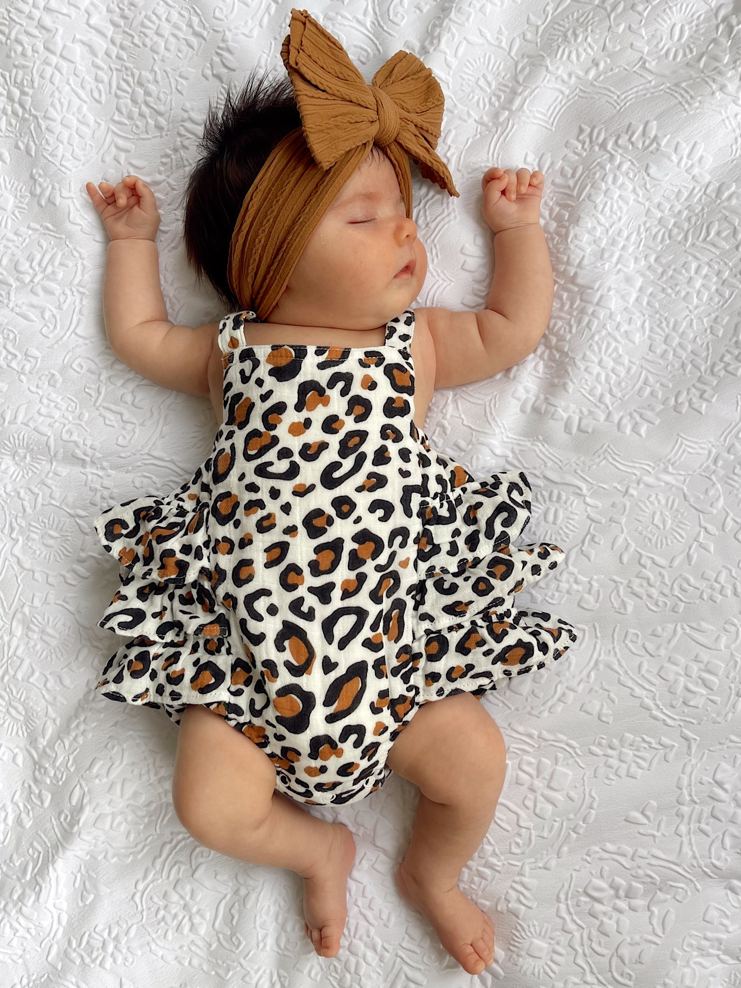 Baby girl sleeps peacefully in a leopard print dress and brown headband on a textured white blanket.