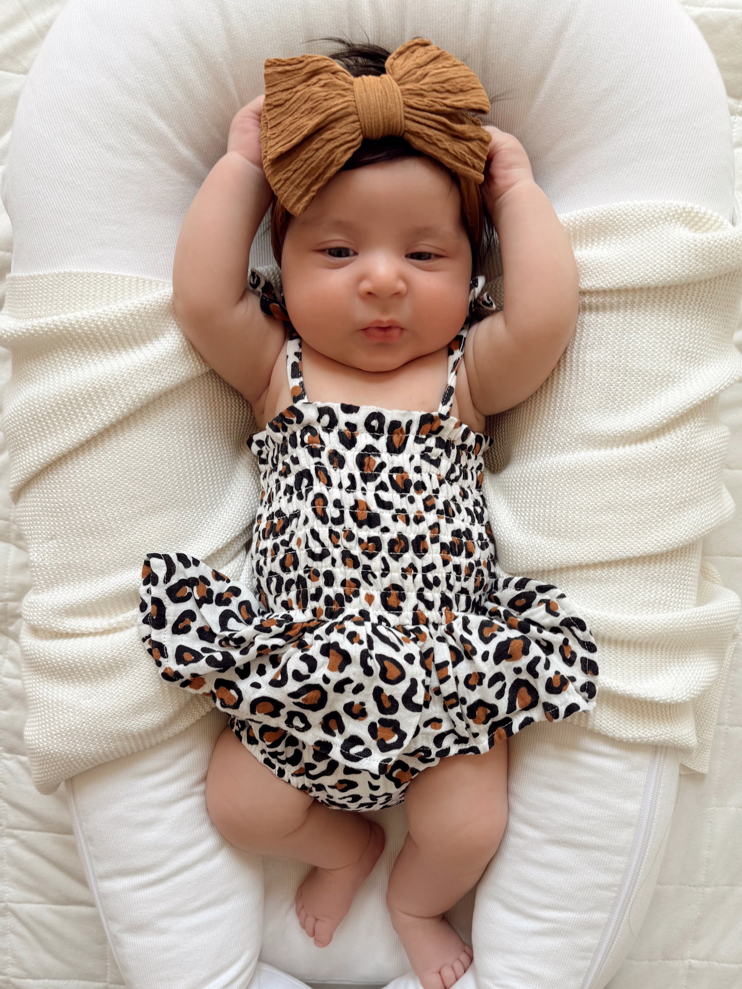 Baby girl in a leopard print dress and a large bow, lying on a cozy white blanket with arms raised.