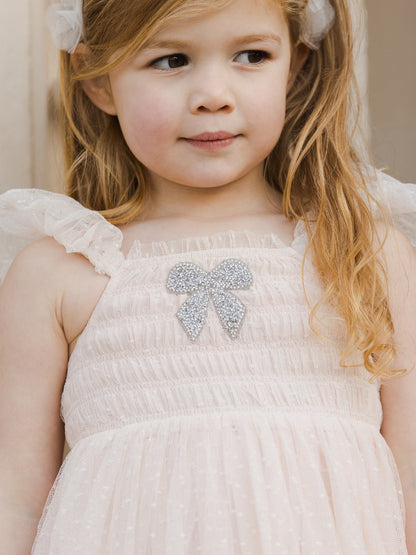 Girl with wavy hair in a pink dress featuring a glittery bow, looking thoughtfully at the camera.