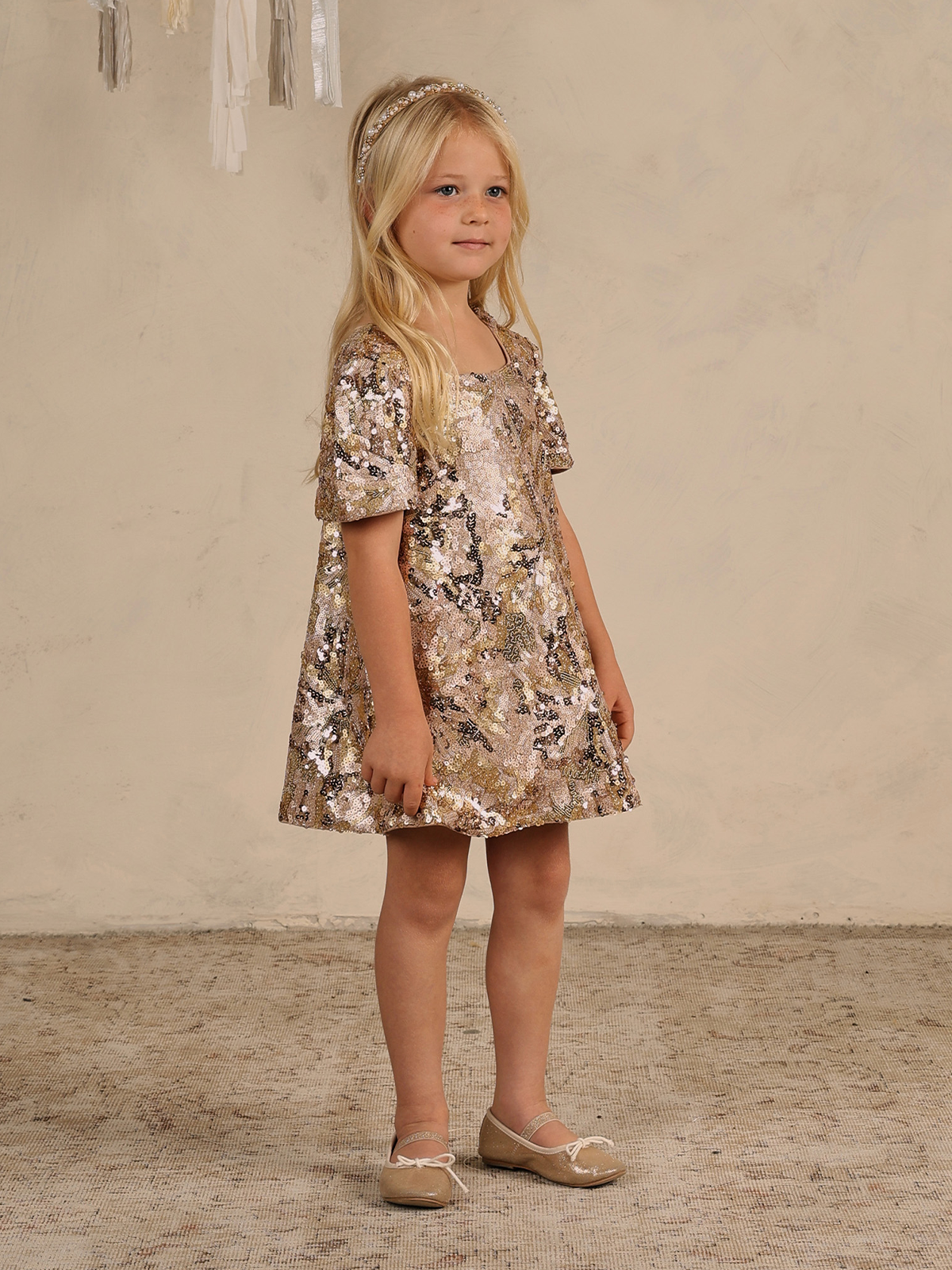 Young girl in a shiny gold sequin dress, standing on a textured floor with a neutral background.