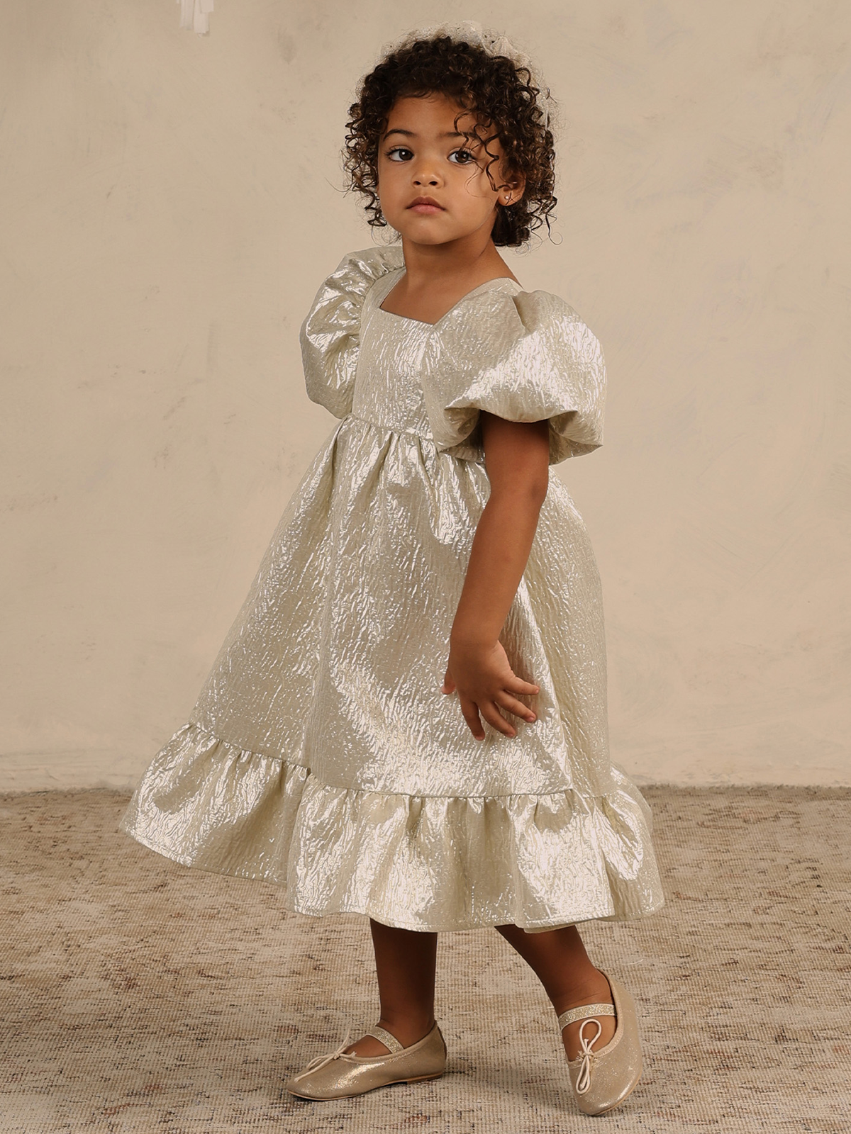 Toddler in a shimmering cream dress with puffed sleeves, standing against a neutral background.