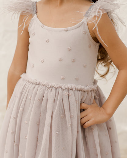 A young girl in a light pink dress with feather straps and floral embellishments, posing confidently with her hand on her hip.