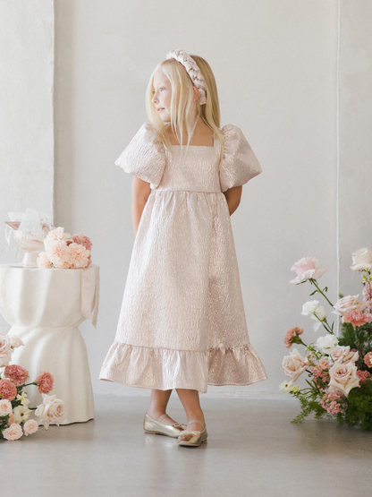 A girl in a pink dress with puffy sleeves stands against a light wall, surrounded by flowers.