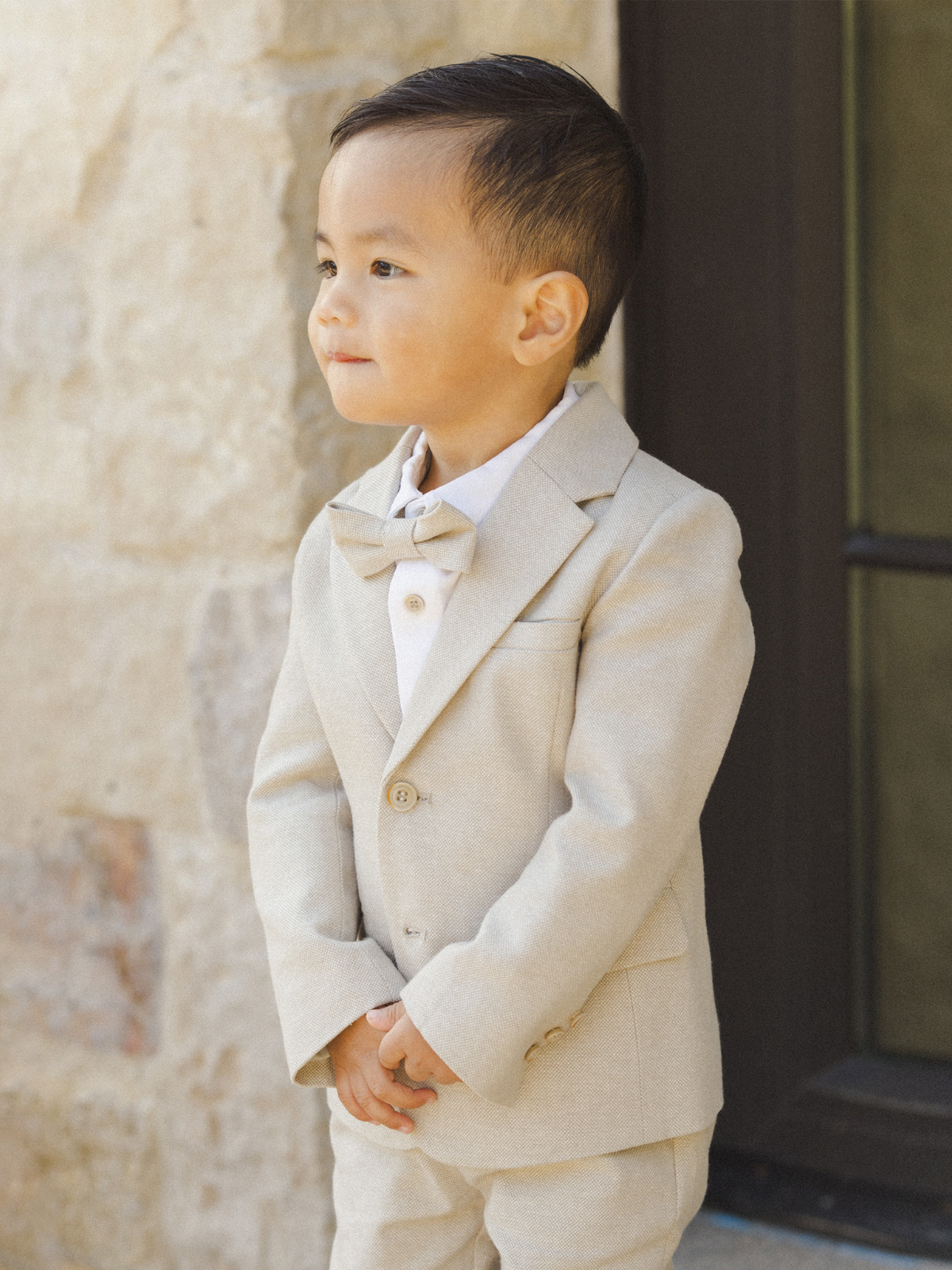 A young boy in a light suit and bow tie stands confidently, hands clasped, against a textured stone wall.