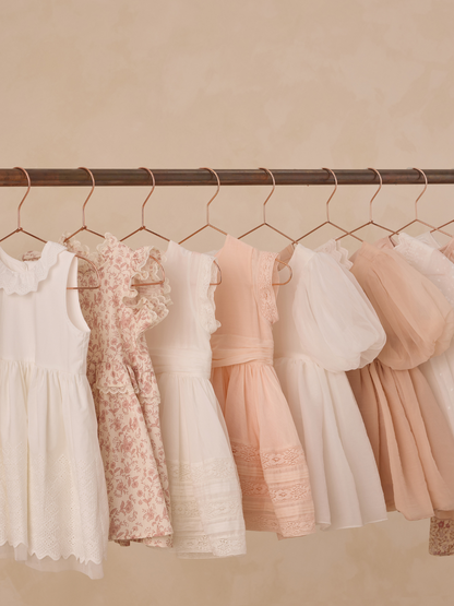 Assortment of children's dresses in soft colors hanging on a copper rack against a beige background.