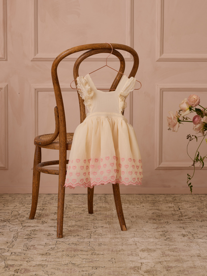 A children's dress with ruffled sleeves hangs on a wooden chair against a soft pink background.