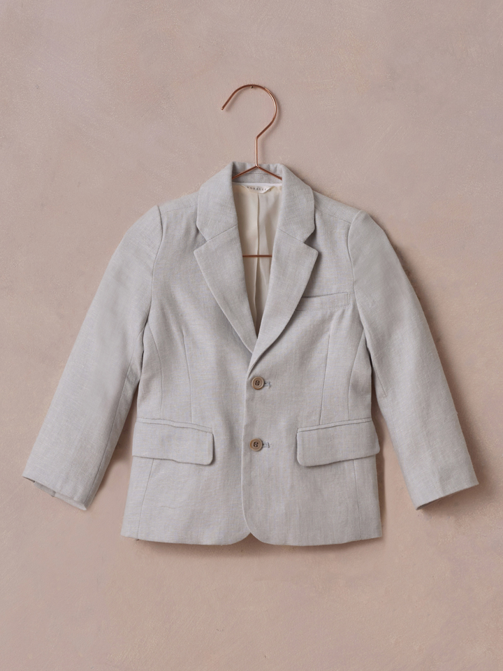 Light gray blazer hanging on a copper hanger against a textured pink background.
