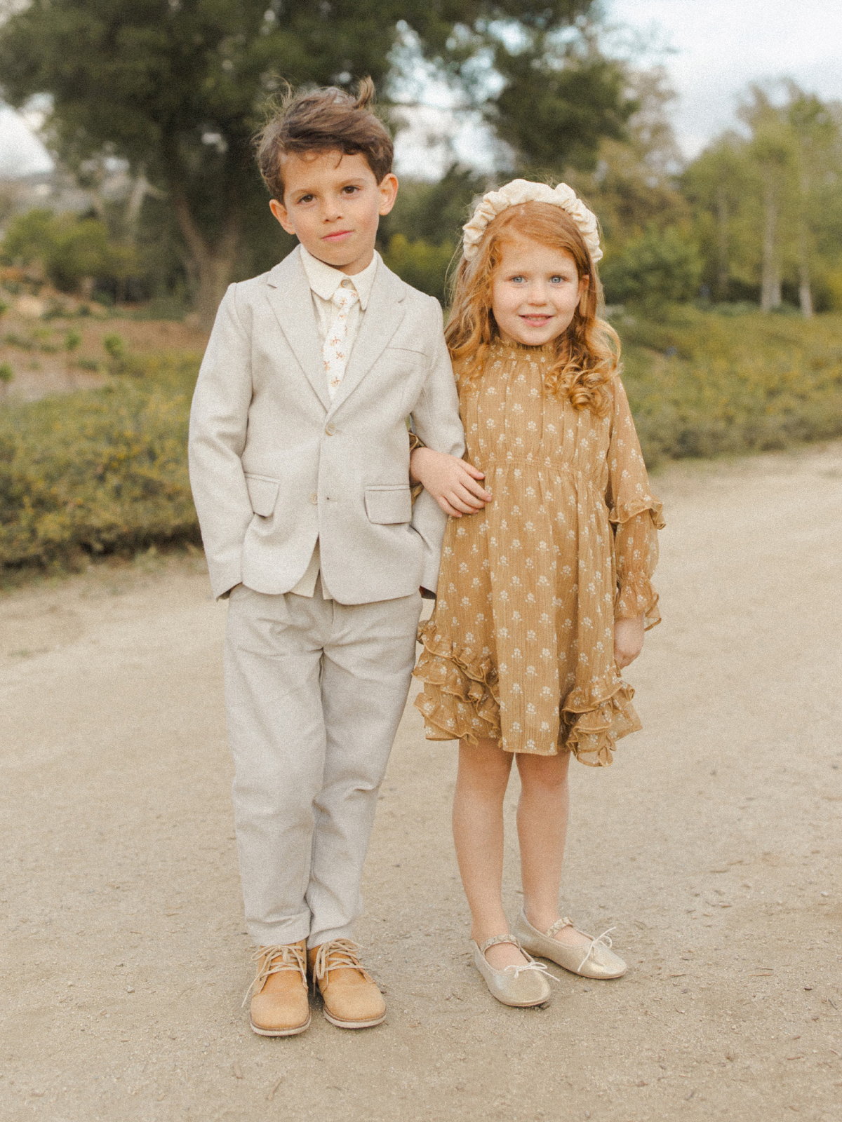 Two children stand together outdoors; a boy in a light suit and a girl in a floral dress and bonnet.