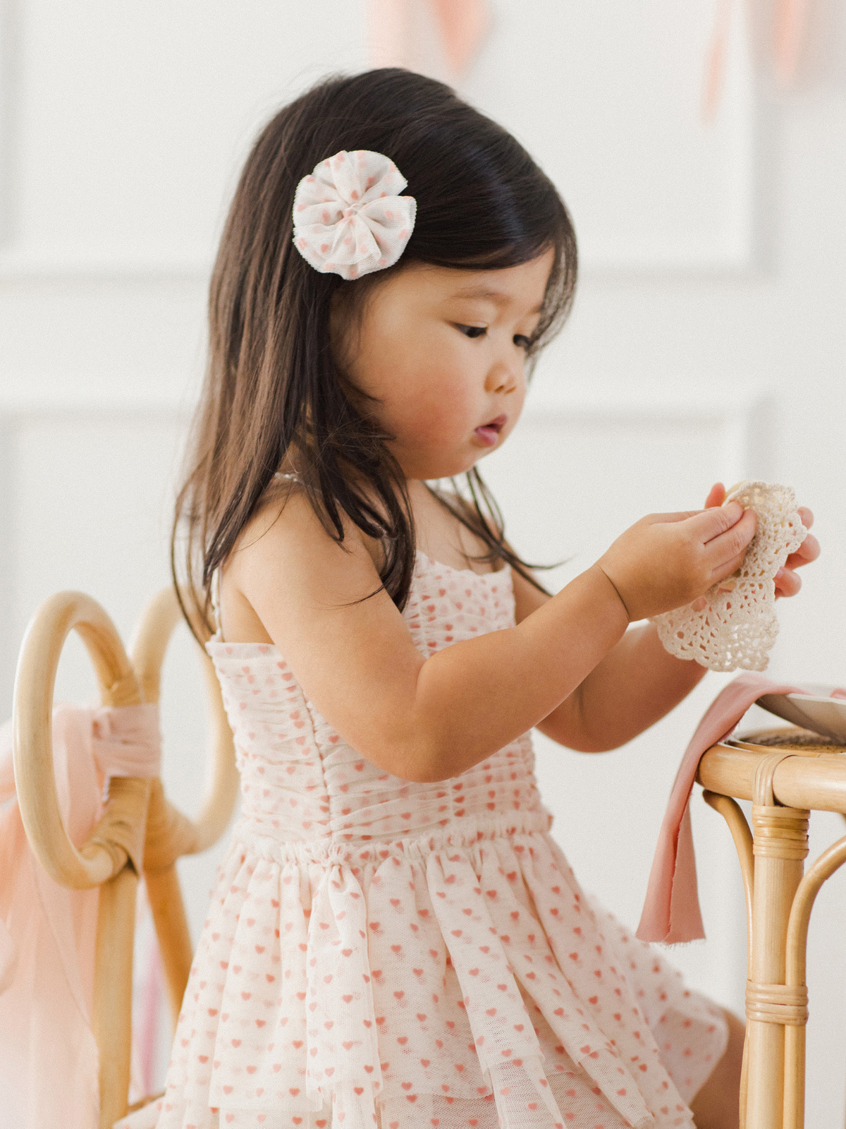 Young girl in a pink heart-print dress, focused on crafting with lace, seated at a light wooden chair.