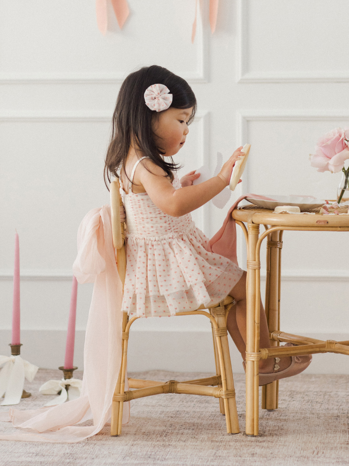 Child in a polka dot dress sits at a table with flowers, holding a cookie amidst soft pastel decor.