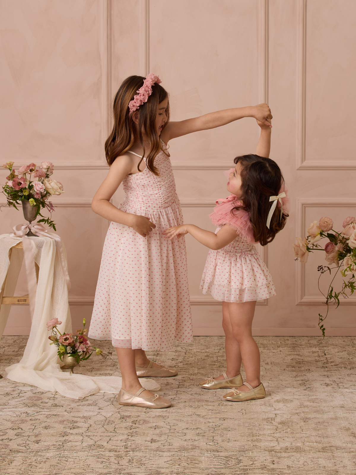 Two girls in pink dresses dance together against a soft pink backdrop, surrounded by floral arrangements.