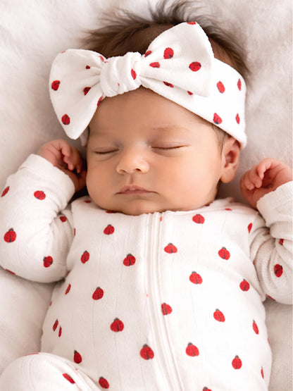 A sleeping baby in a polka dot onesie with a large bow headband, resting on a soft white surface.