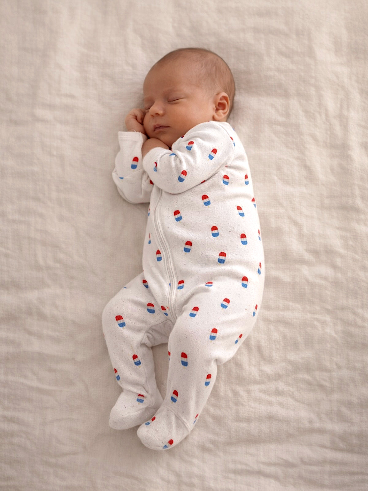 Sleeping baby in a white onesie with colorful patterns, nestled on a soft, light-colored blanket.