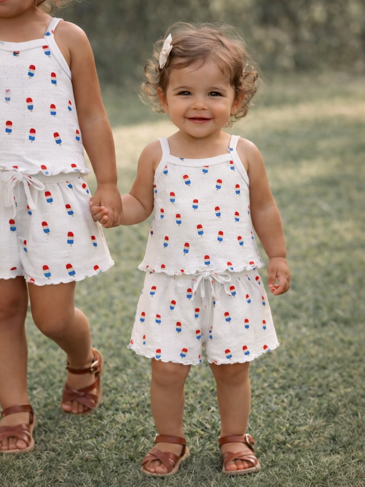 Two young girls in matching playful outfits with popsicle patterns, smiling while holding hands outdoors.