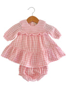Pink gingham dress with long sleeves and ruffled hem, hung on a wooden hanger. Includes matching bloomer bottom.