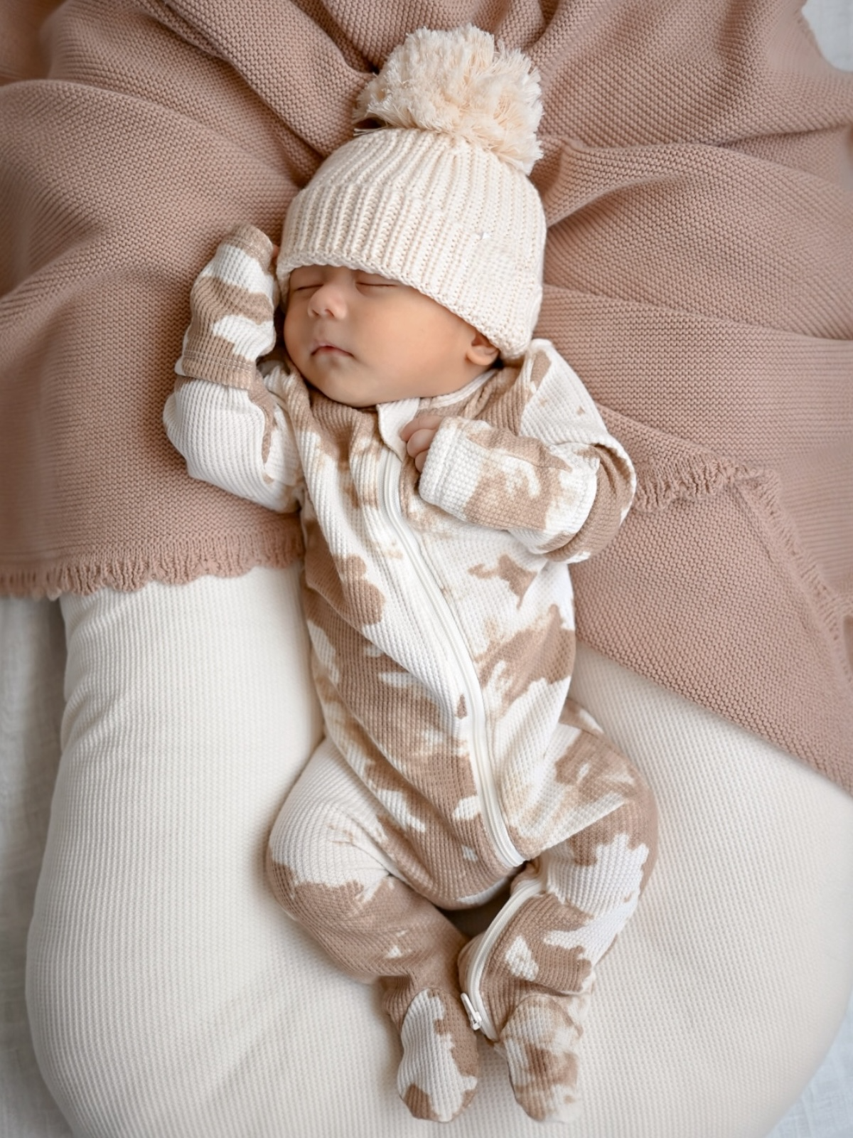 Newborn baby sleeping peacefully in cozy, neutral-toned outfit with pom-pom hat, surrounded by soft fabrics.