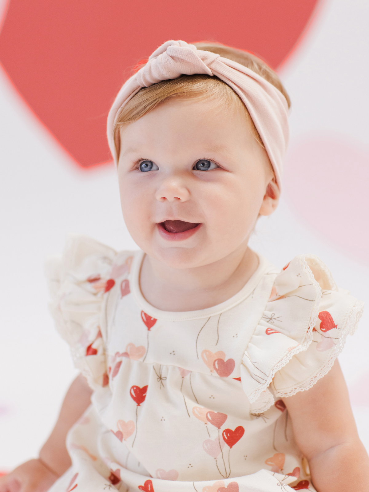 Smiling baby girl in a heart-patterned dress with a pink headband against a soft, colorful background.