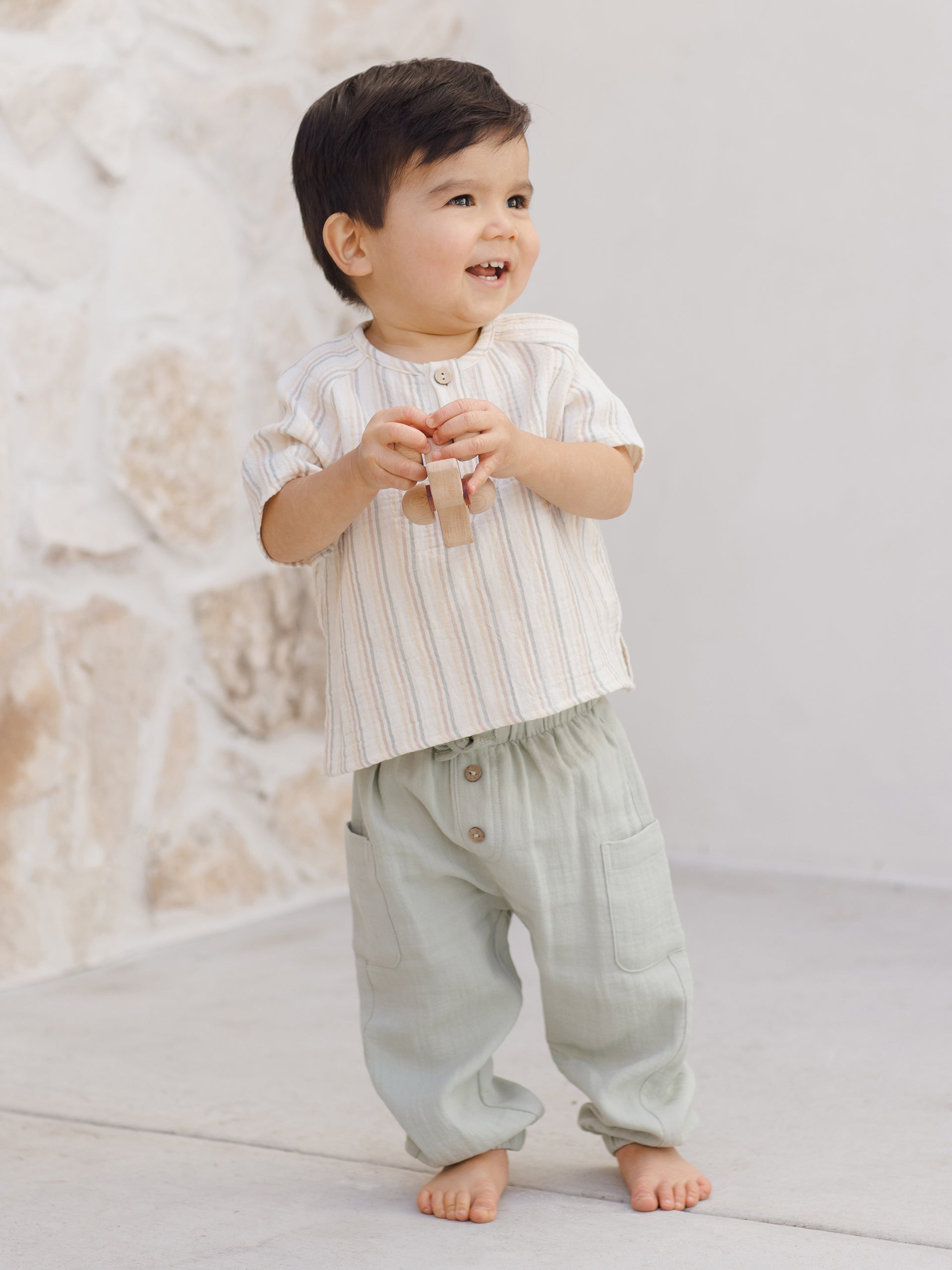 Smiling toddler in a striped shirt and green pants, standing on a light stone surface.