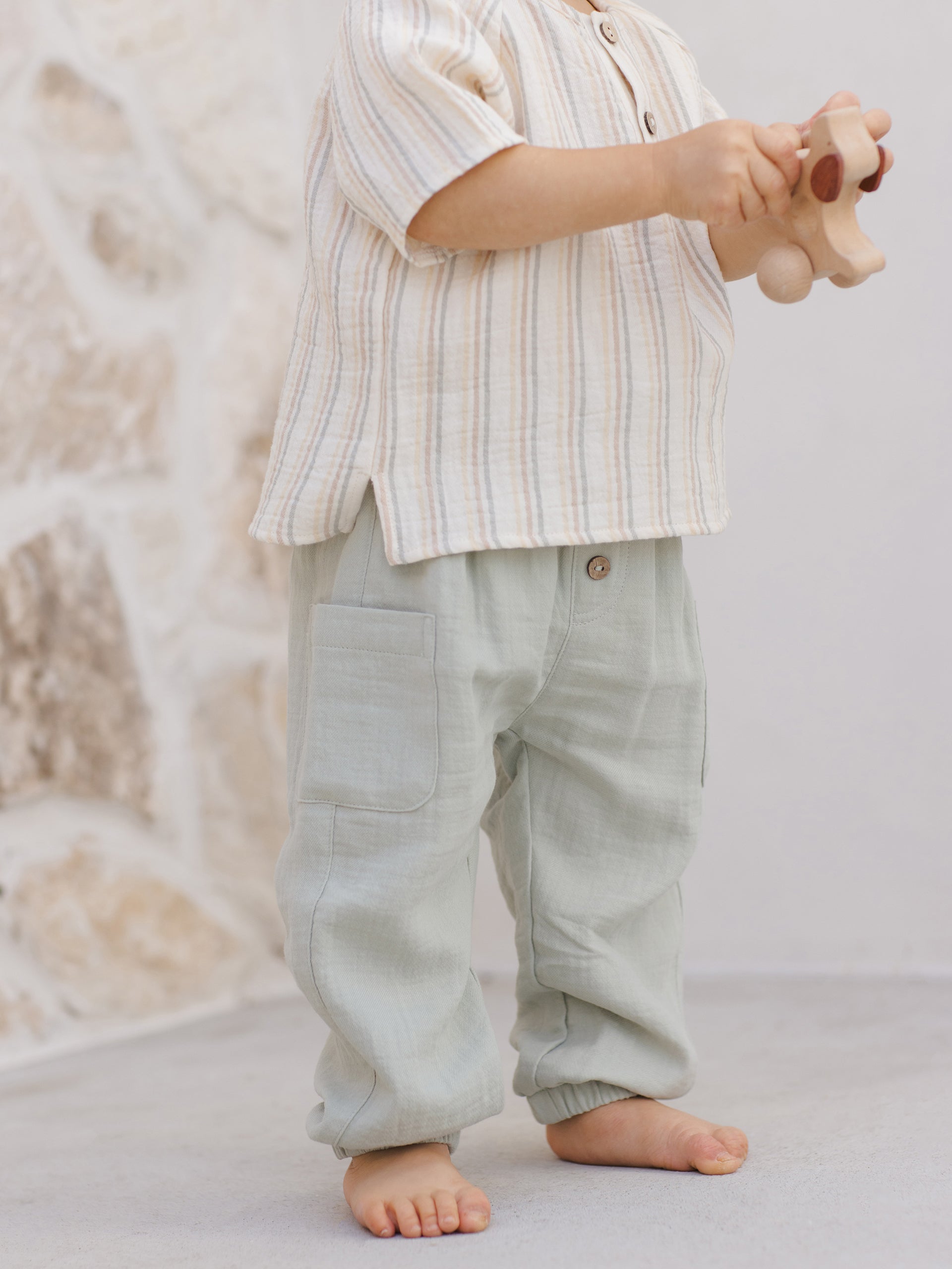 Toddler standing barefoot, holding a toy, wearing a striped shirt and light green pants, on a neutral background.