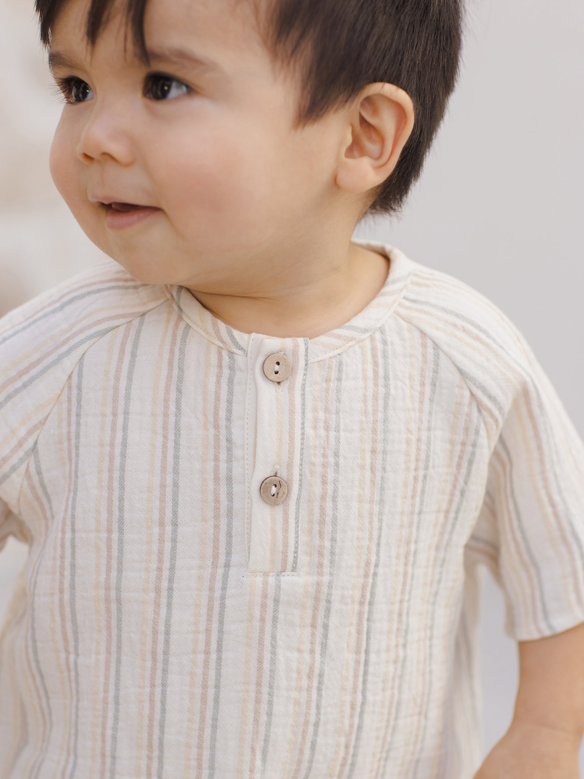 Smiling toddler in a striped shirt, featuring buttons and a relaxed expression, against a neutral background.