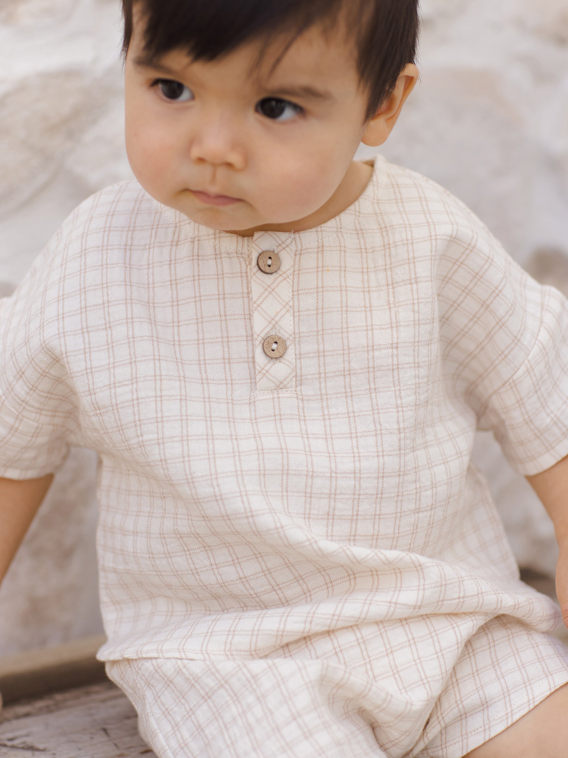 Child in a light plaid outfit with short sleeves, sitting on wooden surface against a textured background.