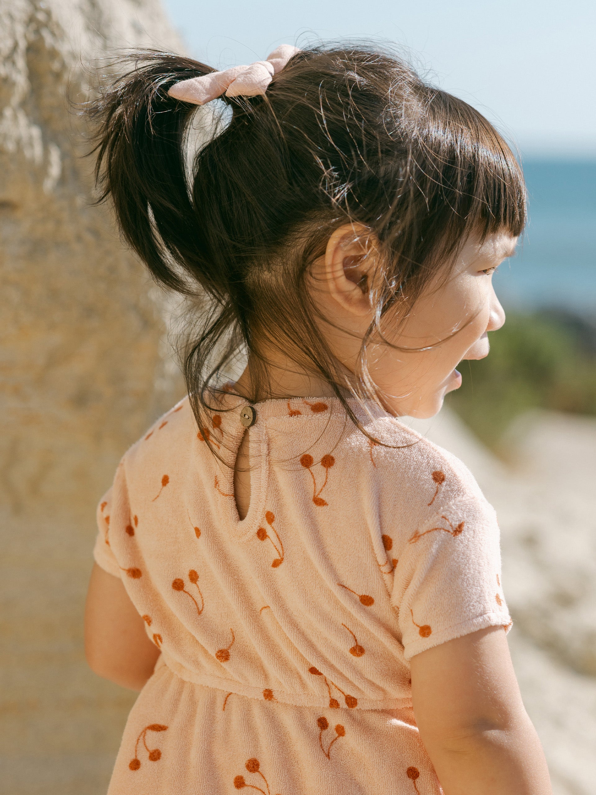 Child with pigtails in a cherry-patterned dress, looking back at a scenic beach view.