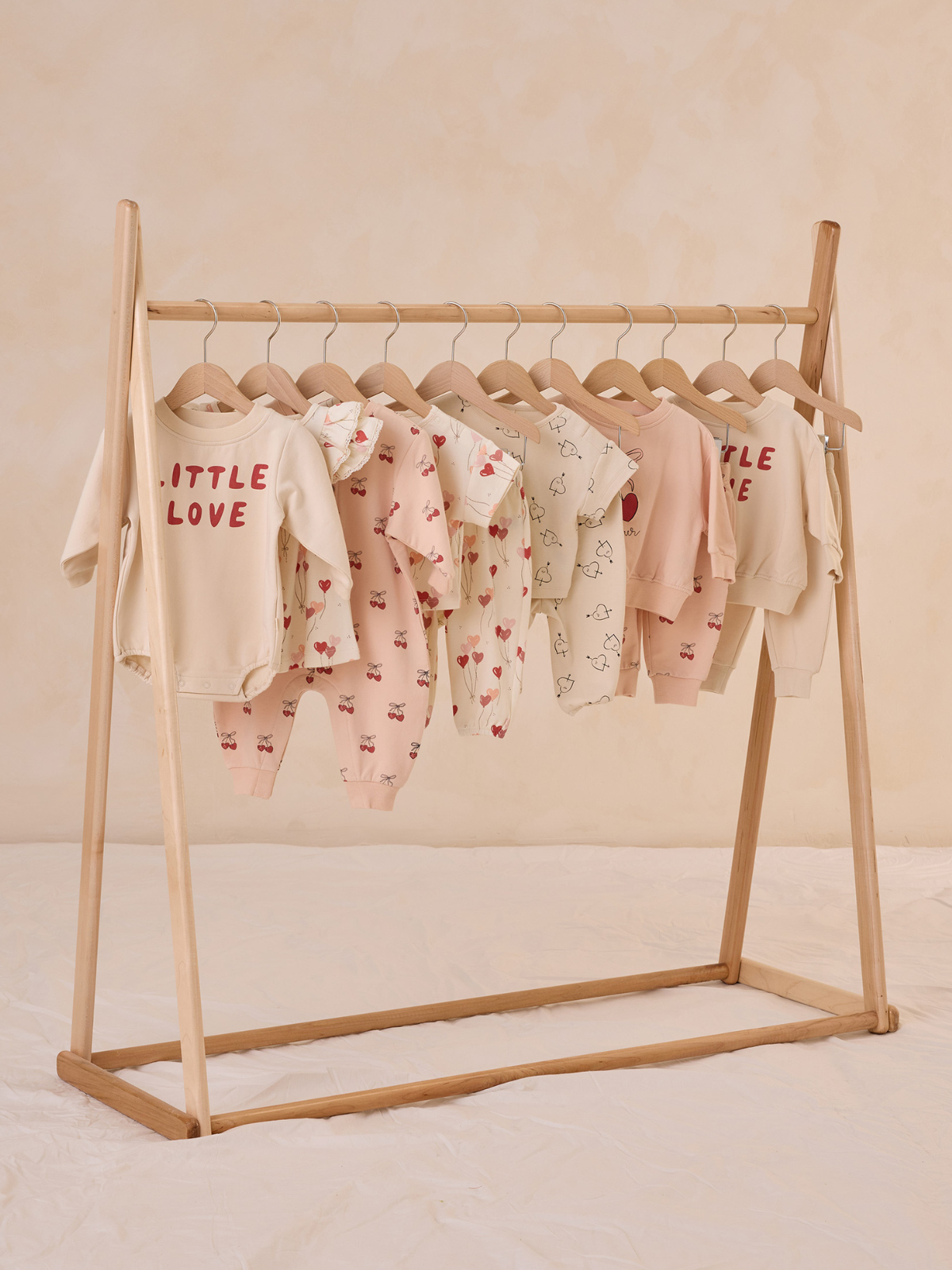 Light-colored baby clothes displayed on a wooden rack against a soft, neutral background.