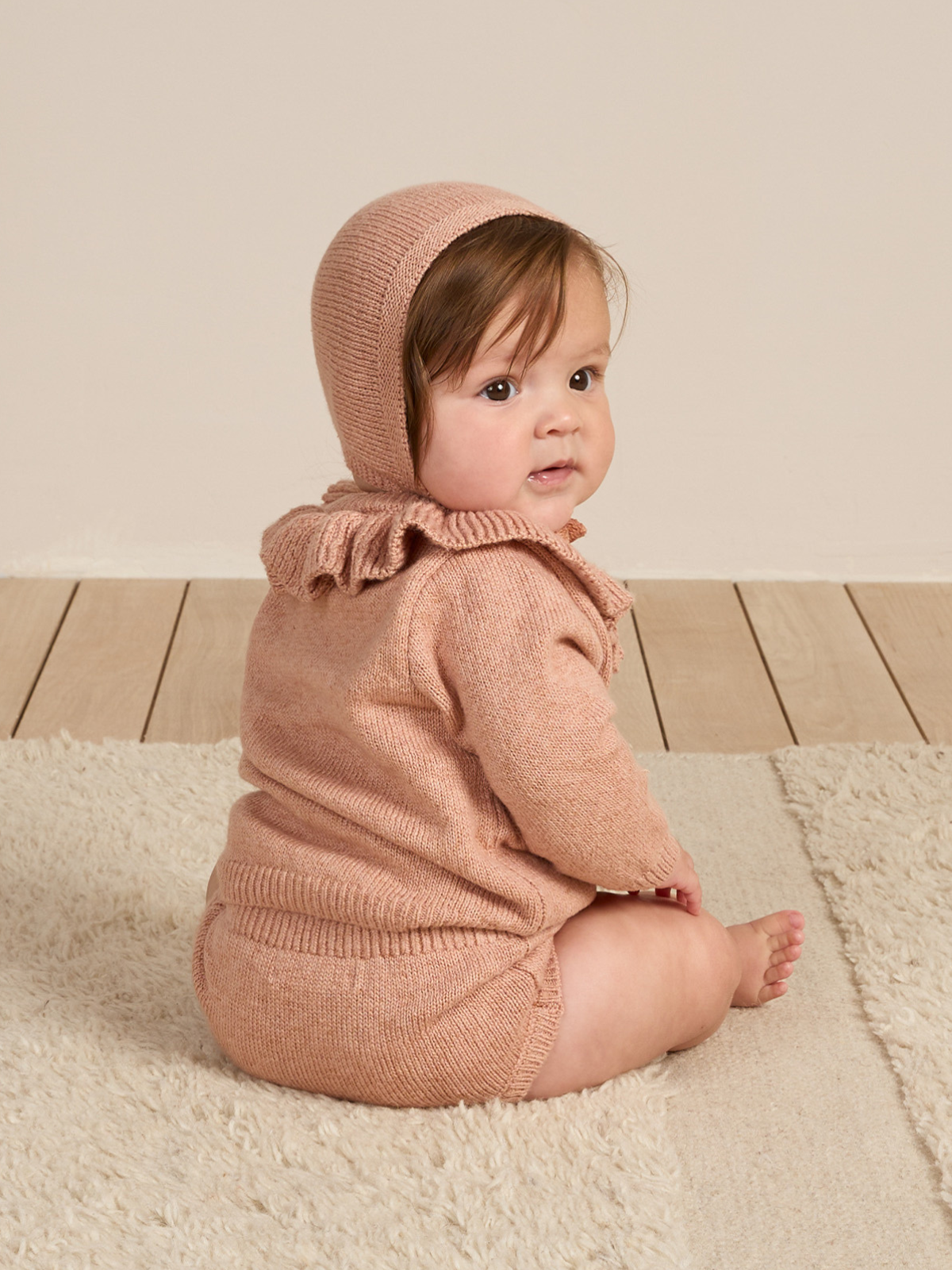 Baby in a soft pink outfit and bonnet, sitting on a plush rug with a wooden floor backdrop.