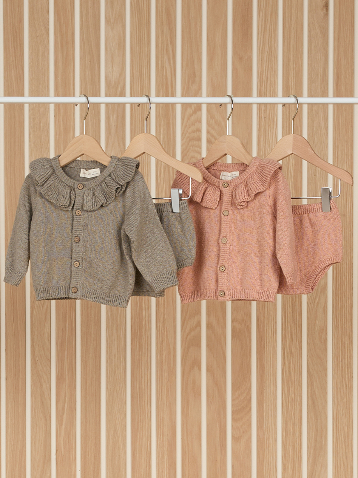 Two baby cardigans with ruffled collars hanging on wooden hangers against a striped wood background.