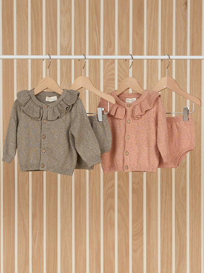 Two baby cardigans with ruffled collars hanging on wooden hangers against a striped wood background.