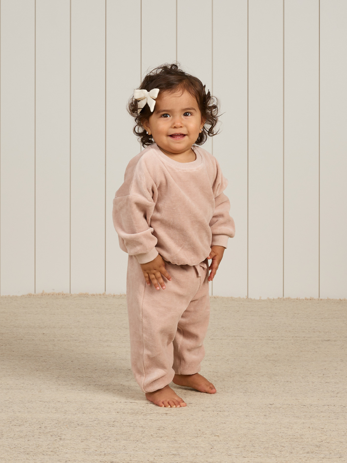 Toddler with curly hair and a bow, wearing a pink outfit, smiling in a light-filled room with wooden accents.