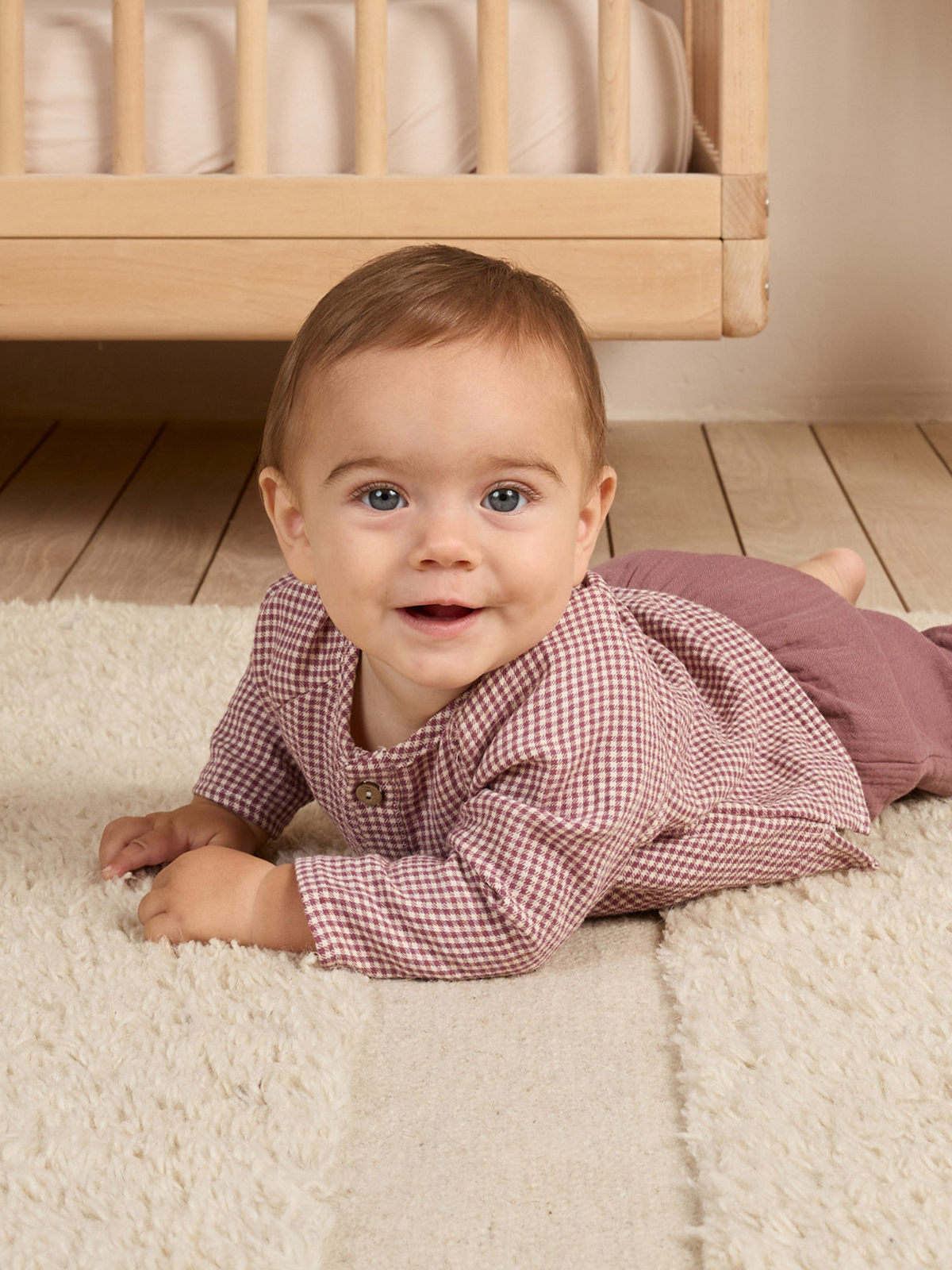 Baby in a checkered outfit smiles while lying on a fluffy rug, with a crib in the background.
