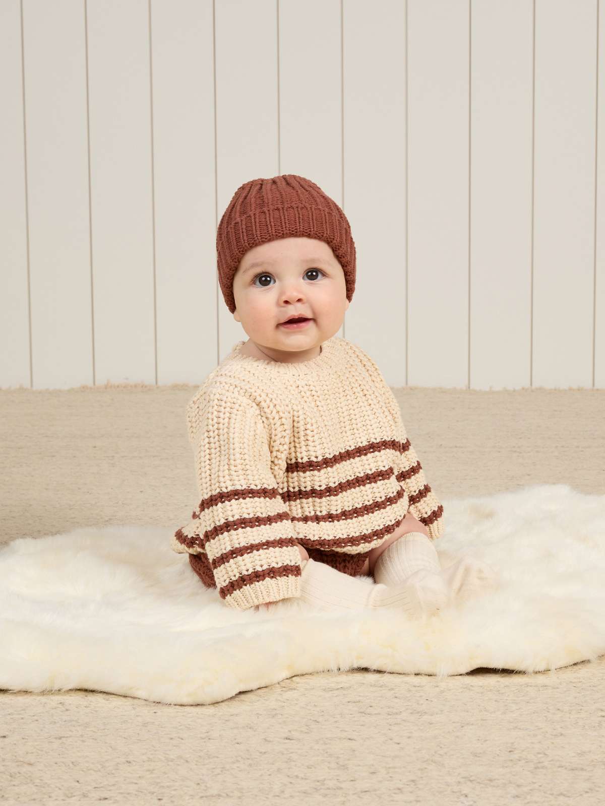 Baby in knitted sweater and hat, sitting on a fluffy rug against a light wooden backdrop.