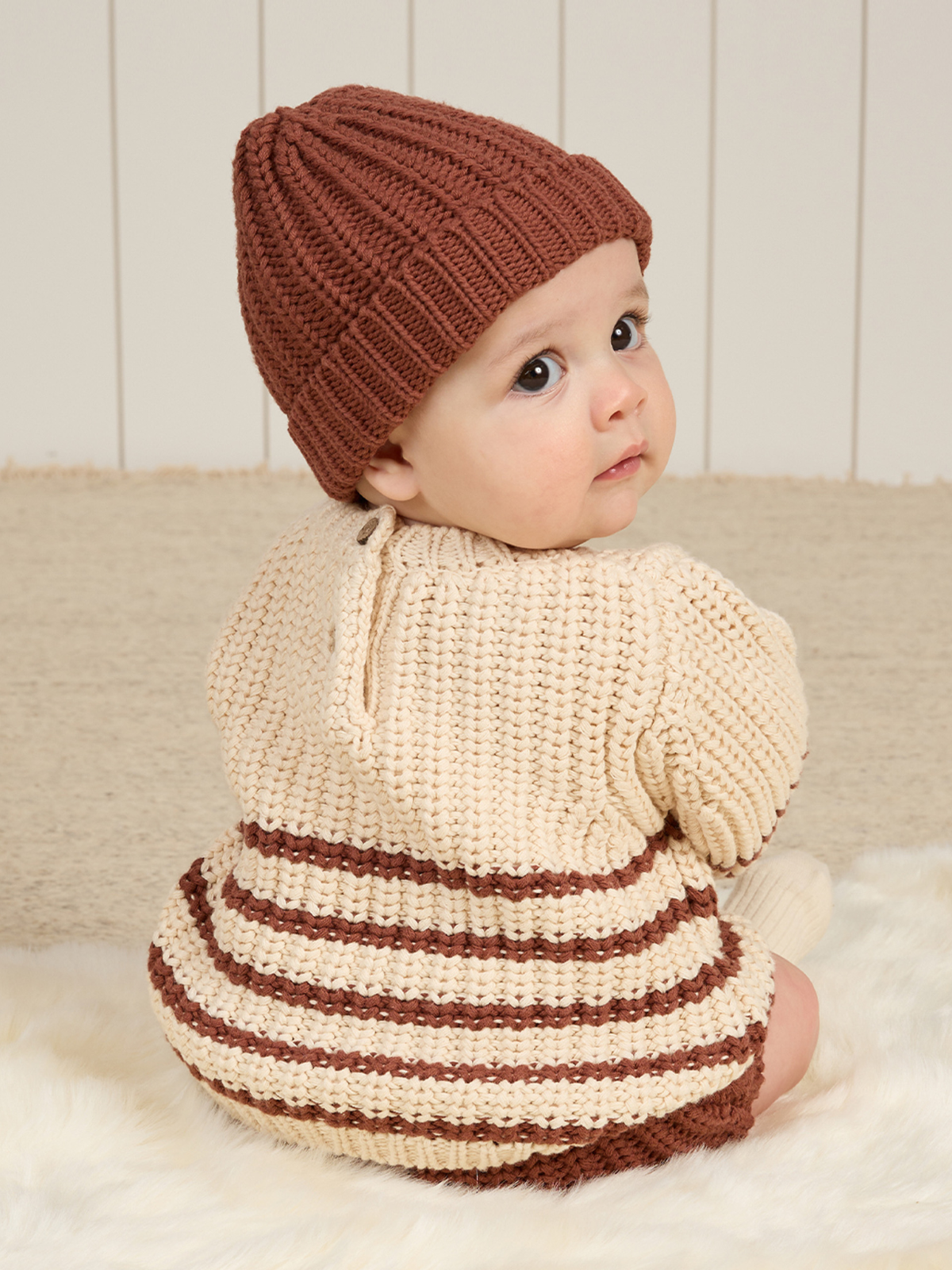 Baby wearing a knitted beige and brown outfit and a brown beanie, sitting on a soft surface, looking back at the camera.