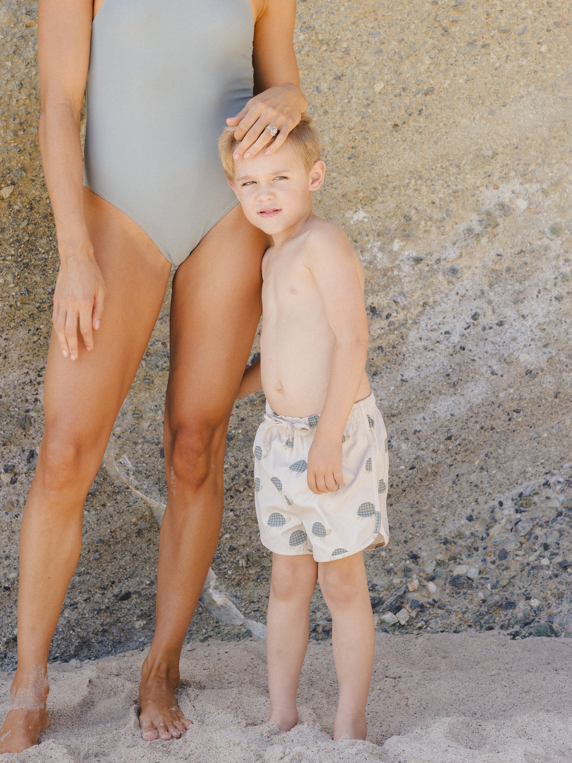 Mother and son standing on the beach, with the mother in a swimsuit and the son in patterned swim shorts.