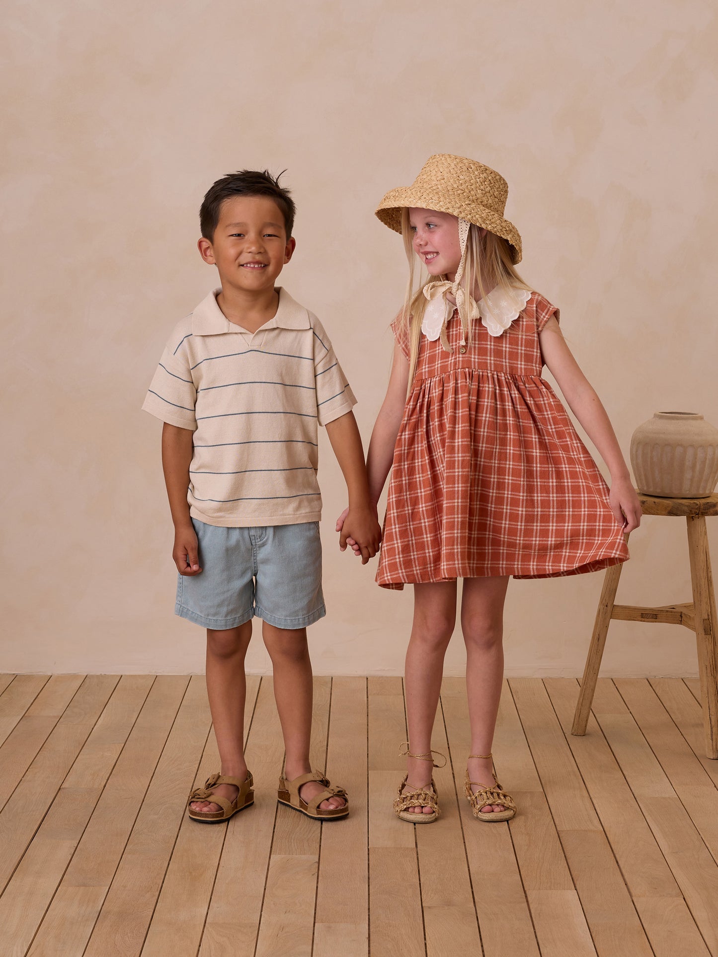 Two children holding hands, smiling. A boy in a striped shirt and shorts, and a girl in a plaid dress with a straw hat.