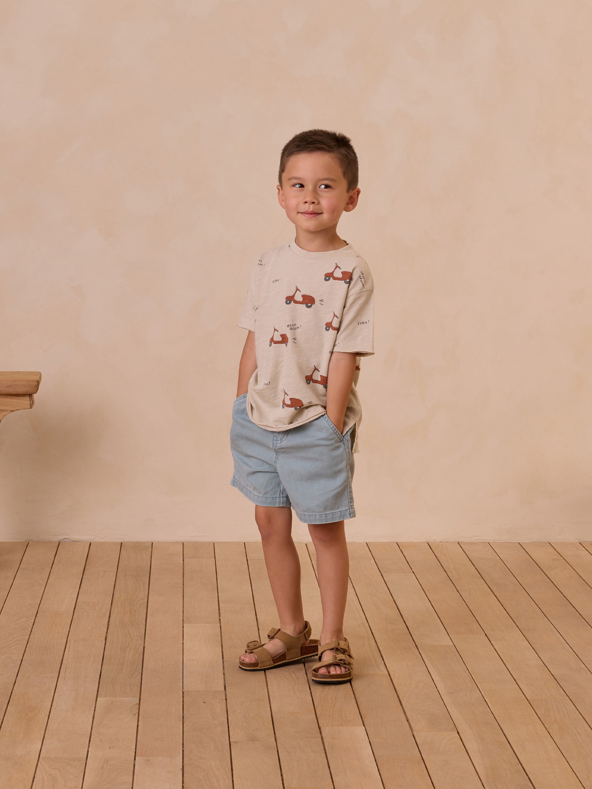 Boy standing with hands in pockets, wearing a printed shirt and shorts, on wooden floor against a beige wall.