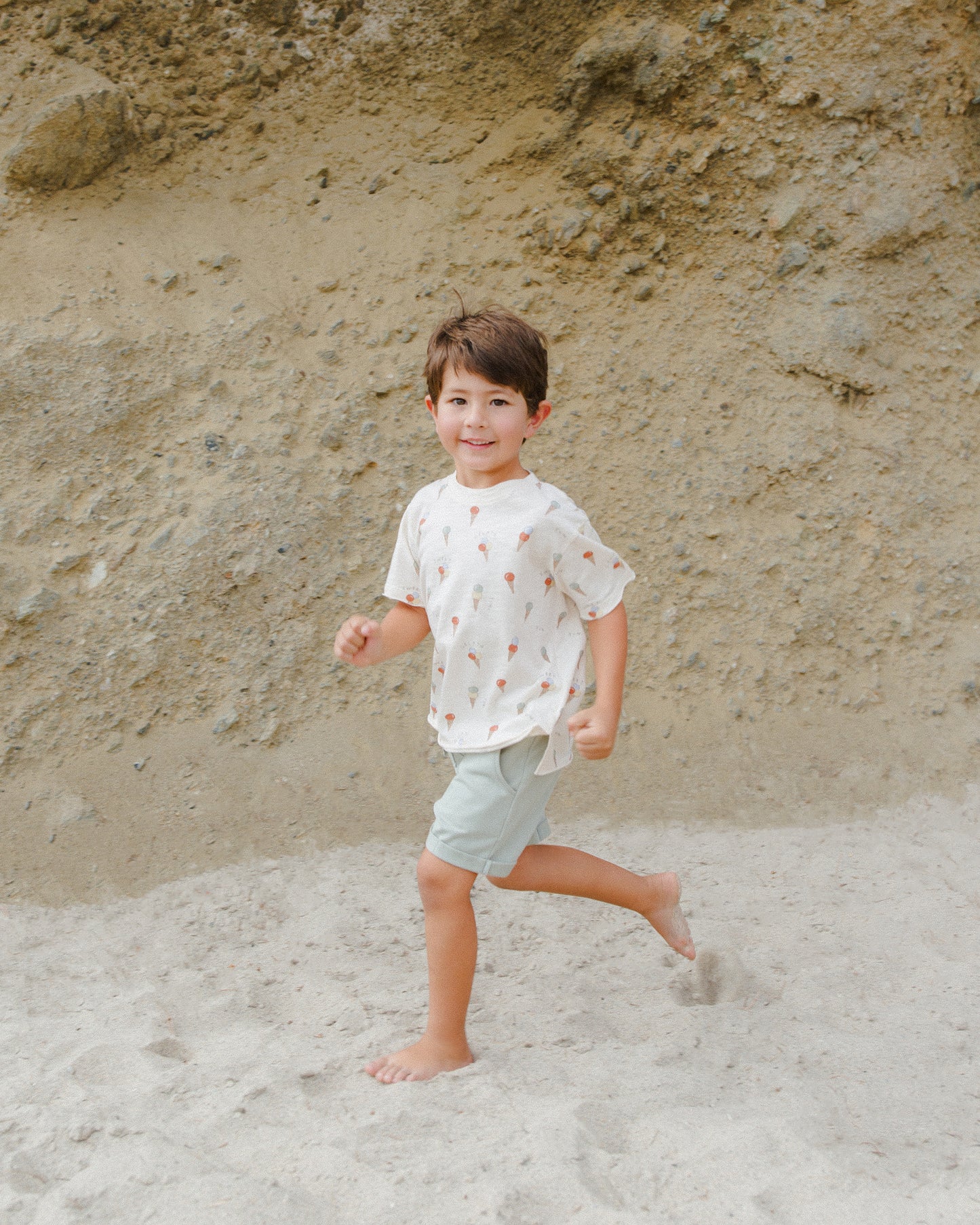 Smiling boy in a playful shirt running on a sandy beach with a rocky background.