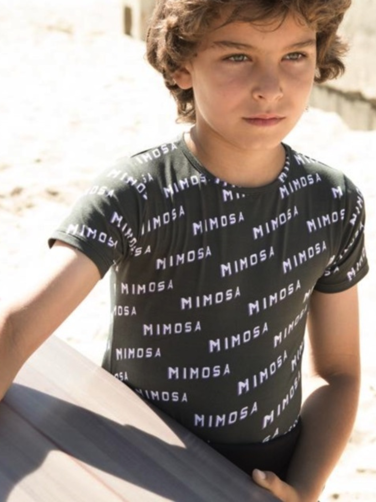 Boy in a patterned T-shirt stands outdoors, holding a surfboard on a sandy beach.