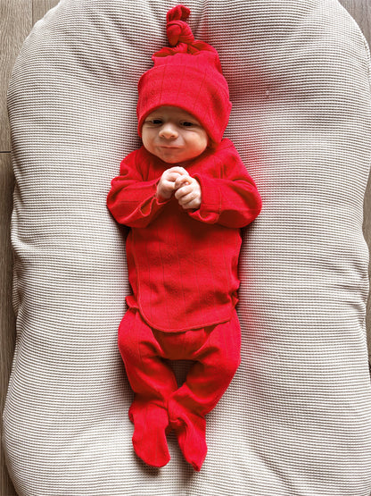 Baby in a red outfit and hat, smiling while lying on a textured cream-colored surface.