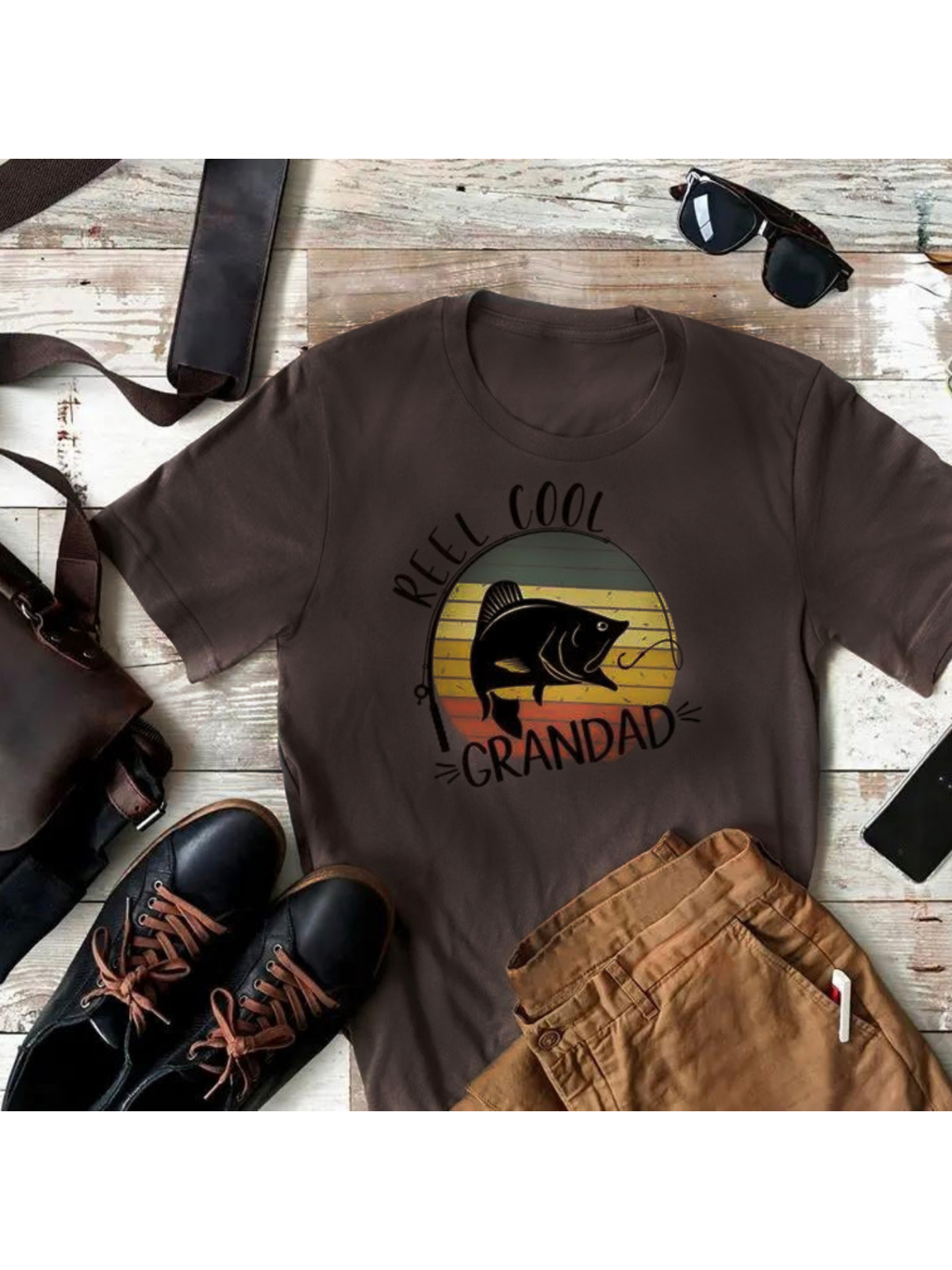 Brown t-shirt with "Reel Cool Grandad" design, fish graphic, surrounded by casual clothing and accessories.