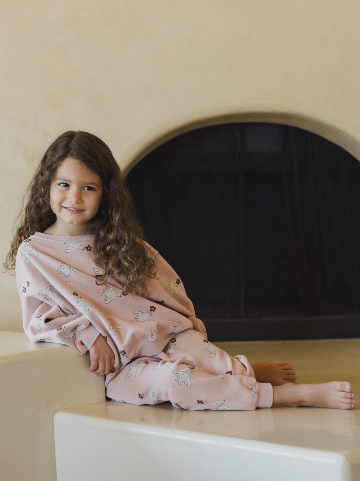 Child with curly hair sitting on a ledge, wearing pink pajamas with a bird print, smiling at the camera.