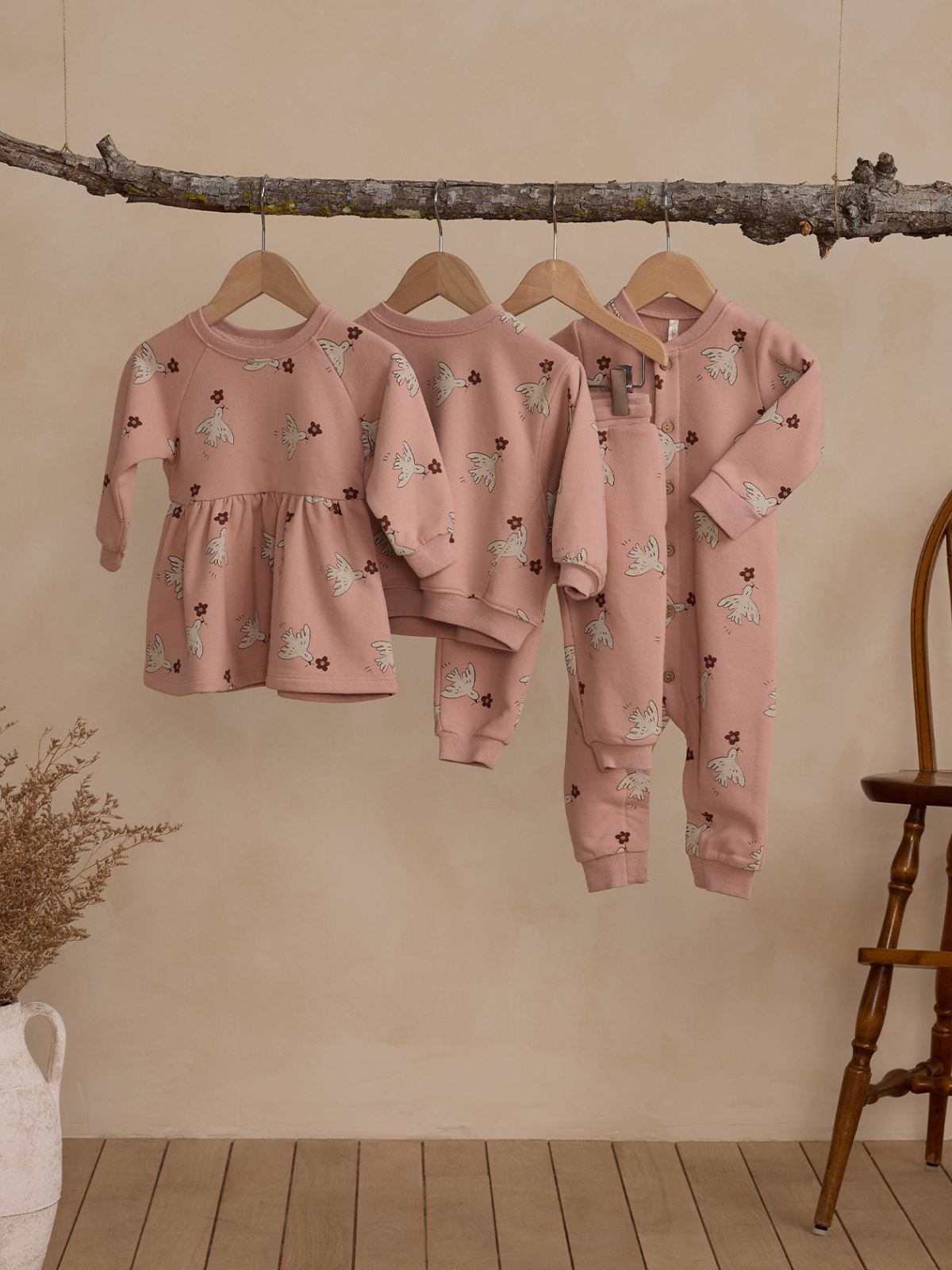 Three pieces of children's clothing in soft pink with floral patterns hanging on a branch against a neutral background.