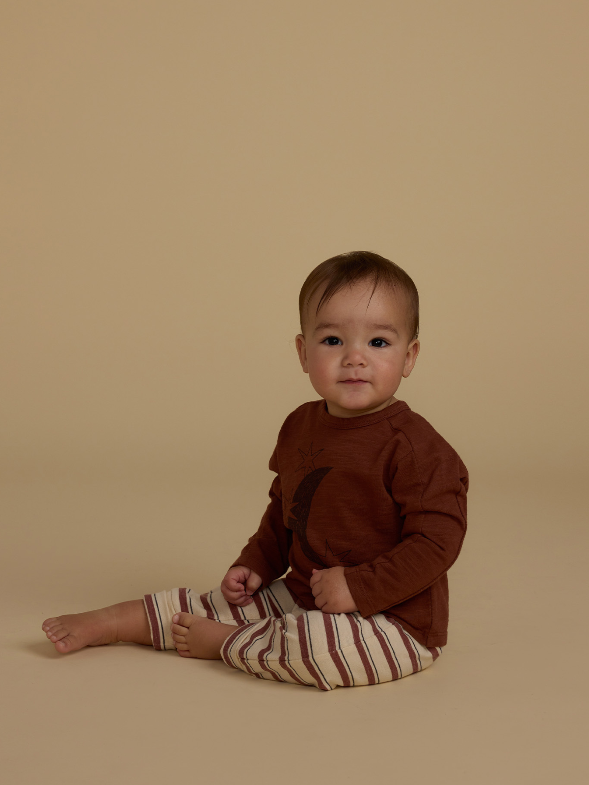 Toddler sitting on a neutral background, wearing a brown shirt and striped pants, smiling gently at the camera.
