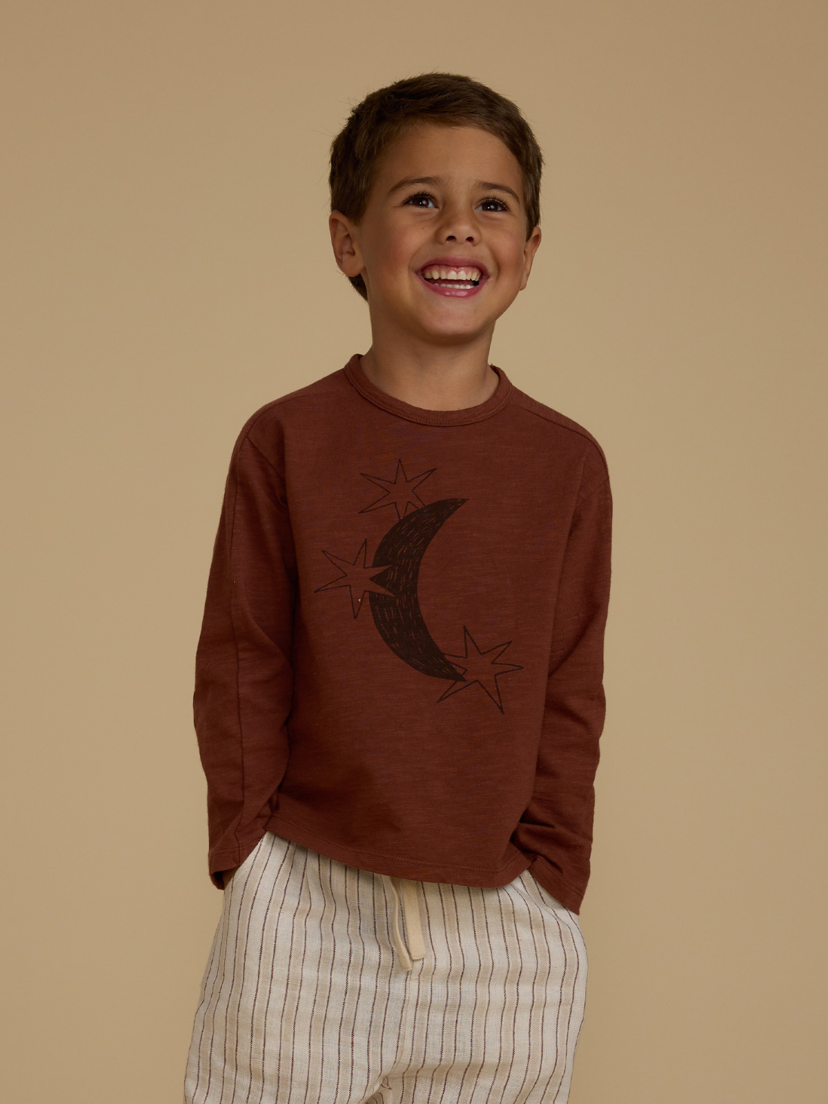 Smiling child in a brown long-sleeve shirt with moon and stars design, wearing light striped shorts against a beige background.